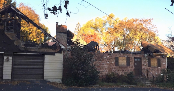 A house with the roof burned off view from the front door showing one garage door off to the left. The windows are boarded up.