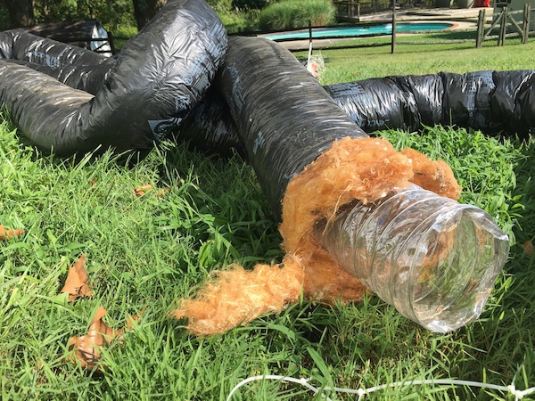 A black flex duct laying out in a grassy yard with orange fiberglass hanging out of the end. There is a swimming pool in the background.