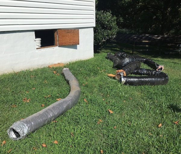 Long tubes of black flex ducts laying out in a grassy yard next to a white house with a crawl space door open to the house.