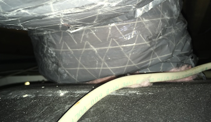 Close up - A gray flex duct with pink fiberglass insulation showing at the end where the duct meets the beam. There is a white wire running past the bottom of the duct and a rip in the casing near the top left of the image.