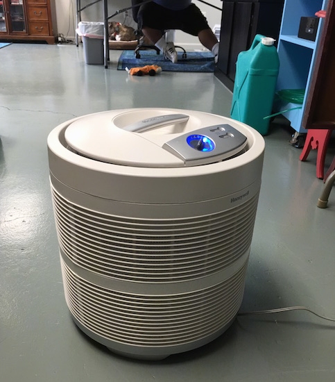 A round white HEPA filter sitting on a gray floor with a person working on a computer and a dog laying down in the distance.