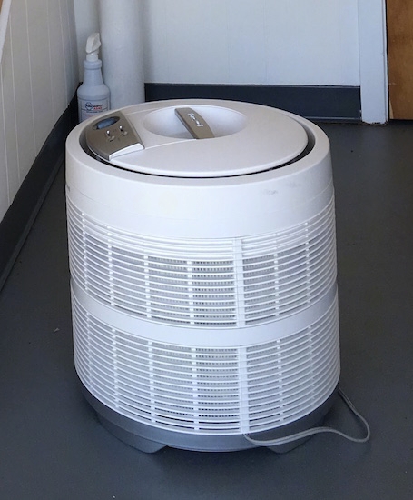 A round white HEPA filter sitting on a gray floor with a bottle of allergy spray in the back corner.