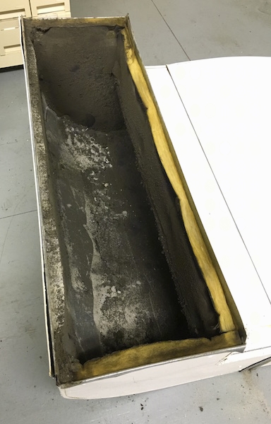 A section of a fiberglass lined duct with lots of black dirt all over the inside laying on a floor after being removed