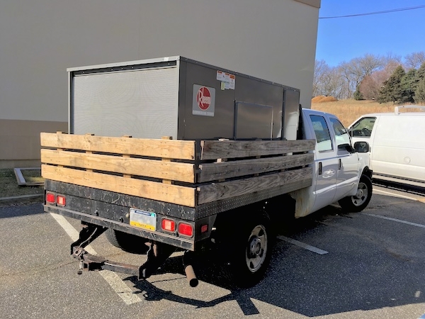 A truck with a large gray HVAC unit in the back bed parked outside of a building.