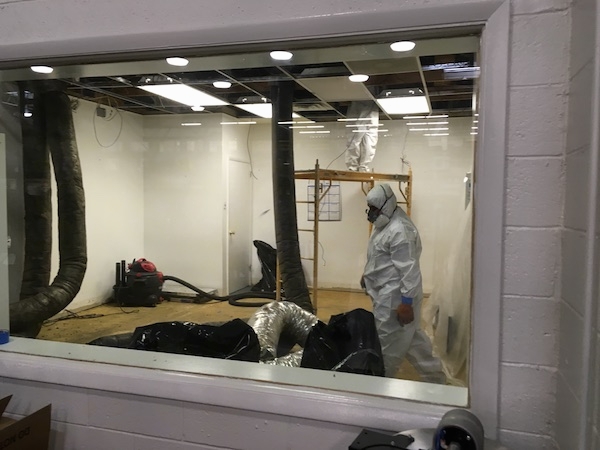 A man wearing a white hazmat suit, a face mask and gloves inside of an offfice while a second man climbs up into the ceiling from scafelting. There are flex ducts all over the floor and hanging from the ceiling.