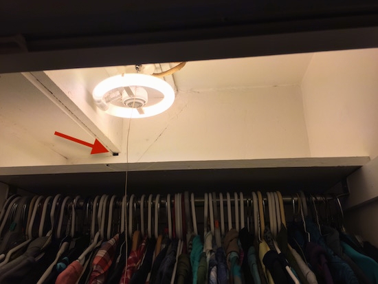 a bedroom closet with clothes hanging up and a red arrow pointing to a hole in the wall above a shelf next to a round fluorescent light