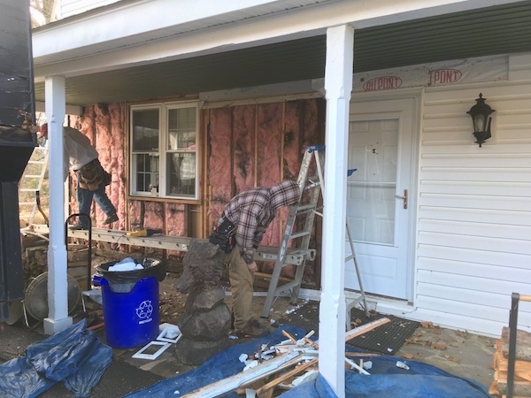 Under a stone porch of an old white farm house with the siding removed from the left half exposing pink fiberglass insulation with black mold lines in it. There are tools and building materials all over.