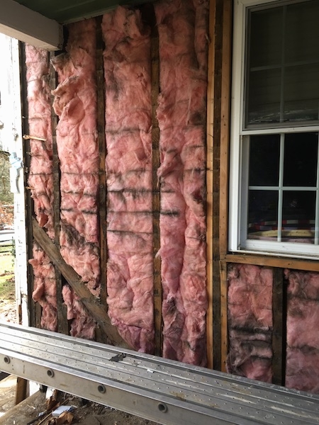 Pink fiberglass insulation on the outside of a house under a porch with black mold and dirt lines in it. There is a window on the right.