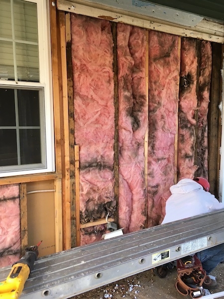 Pink fiberglass insulation on the outside of a house under a porch with black mold and dirt lines in it. There is a window on the left. There is a person in a red hat and gray shirt kneeling down in front of the fiberglass.