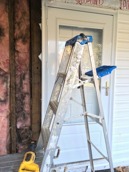 A silver ladder in front of the storm door of a white house under a porch with pink moldy fiberglass off to the left in the wall.