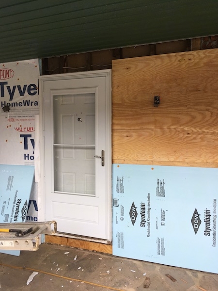 The front of a house with a white door with Tyvek paper on the left and plywood and blue styrafoam on the right. It is under a porch that has a green roof and a stone floor.