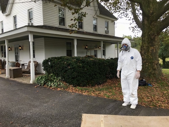 A man in a hazmat suit and gas mask standing in front of a white farm house with a stone wrap around porch.