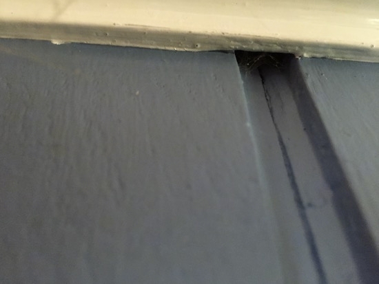 Close up of a seam in a blue painted paneled wall that has a crack where the seam and the white trim meet.