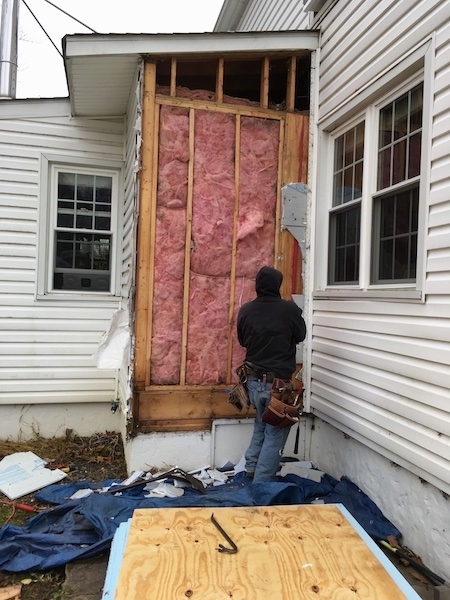 A person in a black sweatshirt standing in front of a white house that has the siding removed exposing the pink fiberglass insulation. There are tools, wood, a tarp and pieces of the house on the ground all around them.