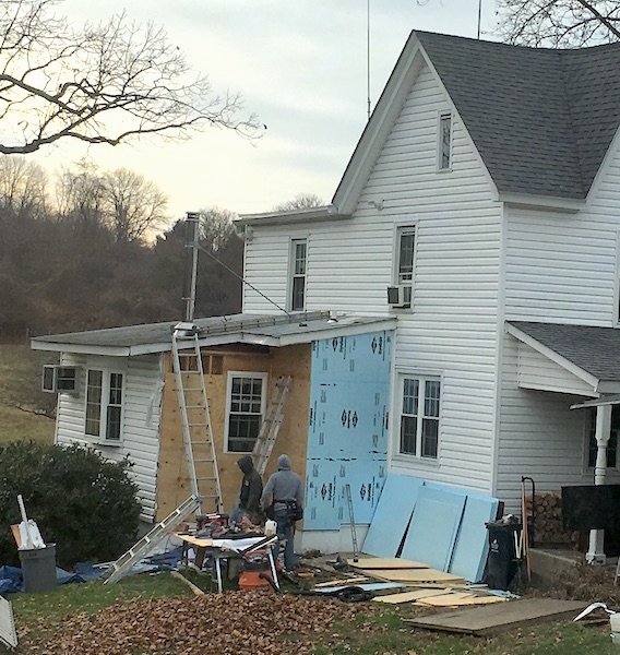 A white farm house with the siding removed from part of the house with wood and blue styrafoam showing with tools and construction materials all around the yard.