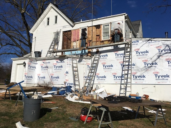 A white farm house with the siding removed on a large part of the house with Tyvek paper showing on the first floor. Up on the second floor are two people removing pink fiberglass insulation from the walls.