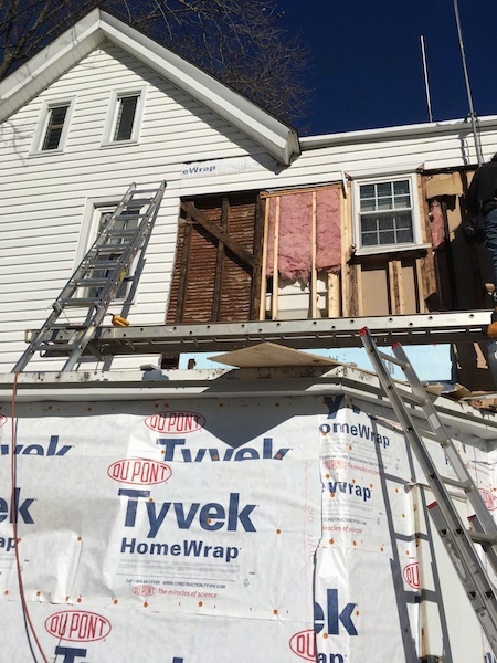 A white house with siding removed showing tyvek paper down low and exposed pink fiberglass upstairs around a window next to lath where the plaster starts. There are two latters leaning against the house.