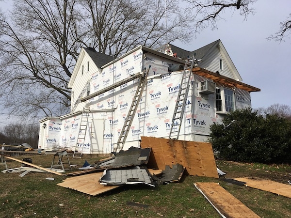 A white house with tyvek paper on the outside, ladders, wood, roof shingles and building materials all over the ground.