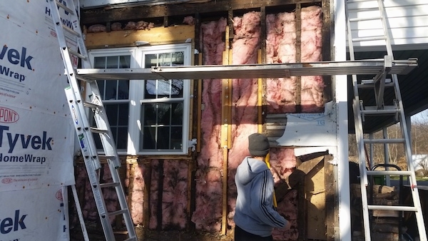 Close up of a white farm house with some of the siding removed showing the pink fiberglass insulation. There is a person dressed in gray standing in front of it.