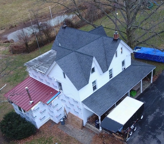 An aerial view of a white farm house with a section of red tin roof on the left and gray shingles on the rest of the house including the wrap around porch. There is a pond behind the house.