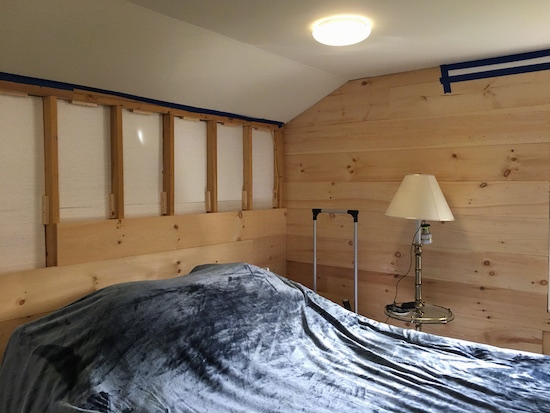 A room under construction being insulated with styrafoam and wood. The on the left is only half way up showing the exposed insulation. There is a blue bed and a lamp in front of the wall.