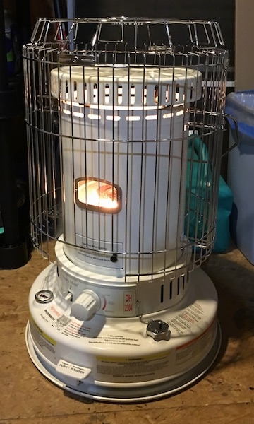 A round white kerosene heater lit in a room that has a wood floor. There is a safety cage built around the heater.