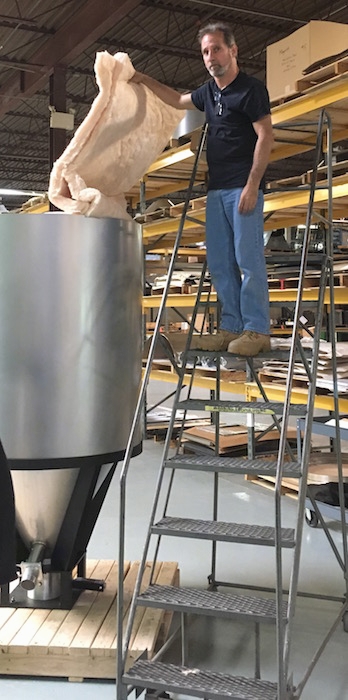 A man standing on a ladder holding up a batt of fiberglass insulation with his bare hand and wearing no mask over a metal tube in a shop.