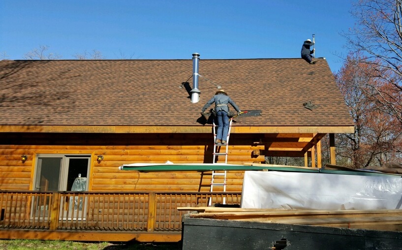 A wooden log cabin with a wrap around deck. There is a dumpster in front of it, and a man on a ladder about to climb up on the roof with a second man at the top peak of the roof.