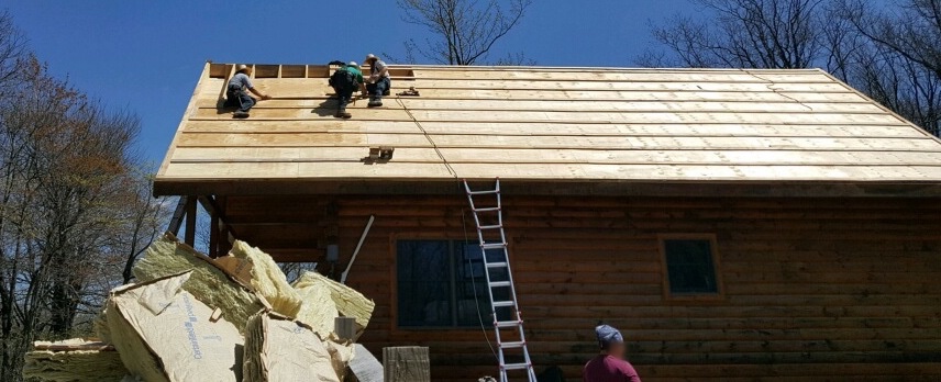 Three men up on the roof of a wooden log cabin putting down new plywood. There is a fourth man standing on the ground next to a very large 8 foot pile of yellow fiberglass batts.