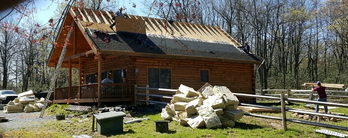 Front side view of a wooden log cabin with shingles remaining on the lower end of the roof. They were removed from the top near the peak and there is a large pile of fiberglass on the ground in front of the cabin and a second pile on the side of the cabin.