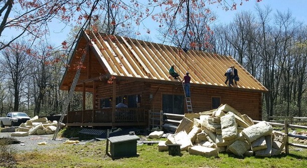 The front side view of a wooden log cabin with four men up on the roof after the shingles and plywood had been removed. There is a very large pile of fiberglass on the side of the cabin with a second pile in front of the cabin.