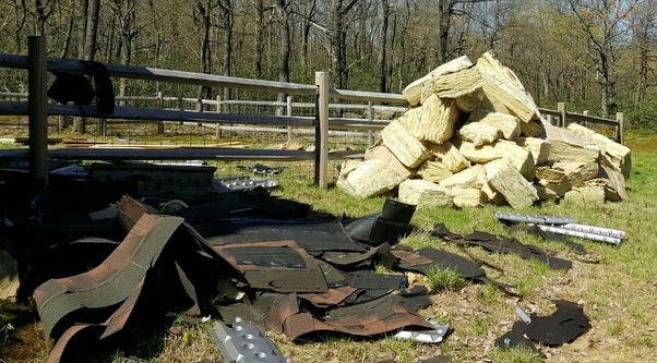 A large pile of thick yellow fiberglass batts laying on the ground in front of a pile of black roof shingles in front of a split rail fence.