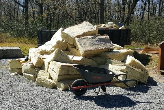 A large pile of yellow fiberglass batts laying in the stone gravel driveway with a wheel barrow sitting in front of it.