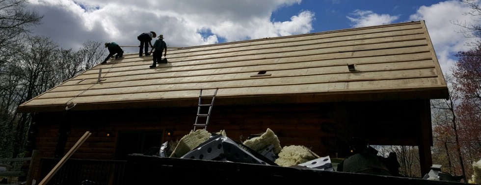 Three men on top of a log cabin roof nailing up new plywood. There is a large dumpster full of yellow fiberglass batts in front of them.