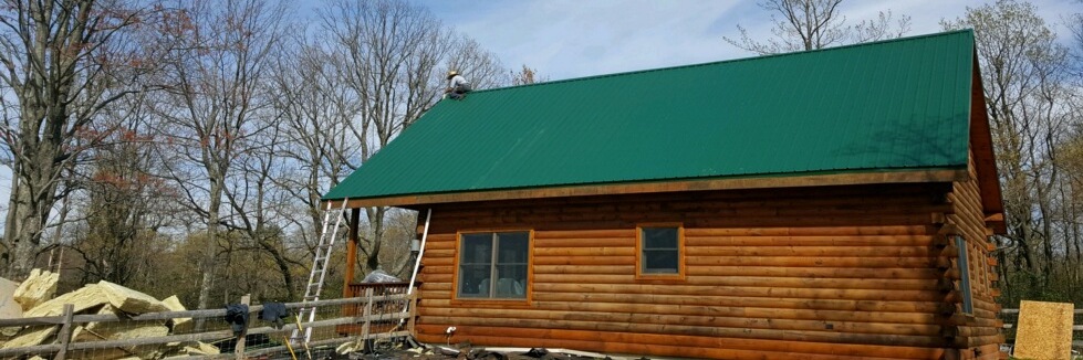 The side of a log cabin with a green tin roof. There is a man on the top of the peak of the roof and a pile of yellow fiberglass on the ground.