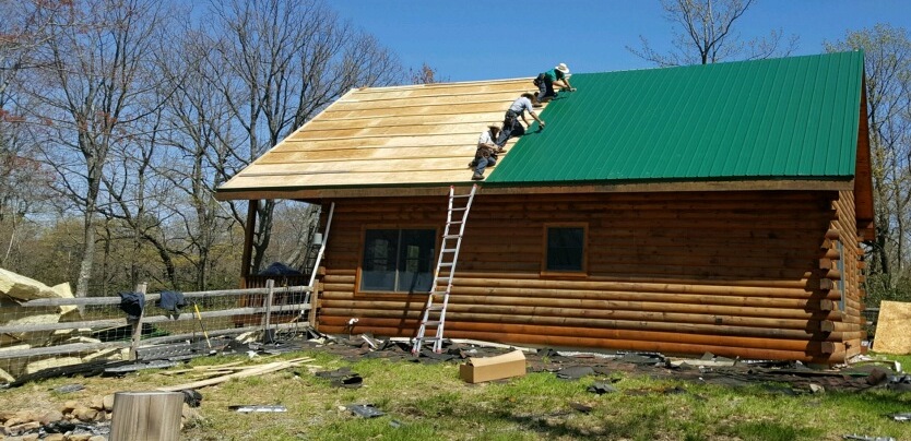 A wooden log cabin with half of the roof green tin and the other half plywood. There are four men on the roof laying down the green tin and a pile of yellow fiberglass on the left of the image on the ground. The cabin has a split rail fence around the back yard.