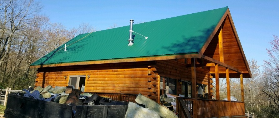 Front side view of a wooden log cabin with a green tin roof. There is a dumpster full of yellow fiberglass batts in front of it.