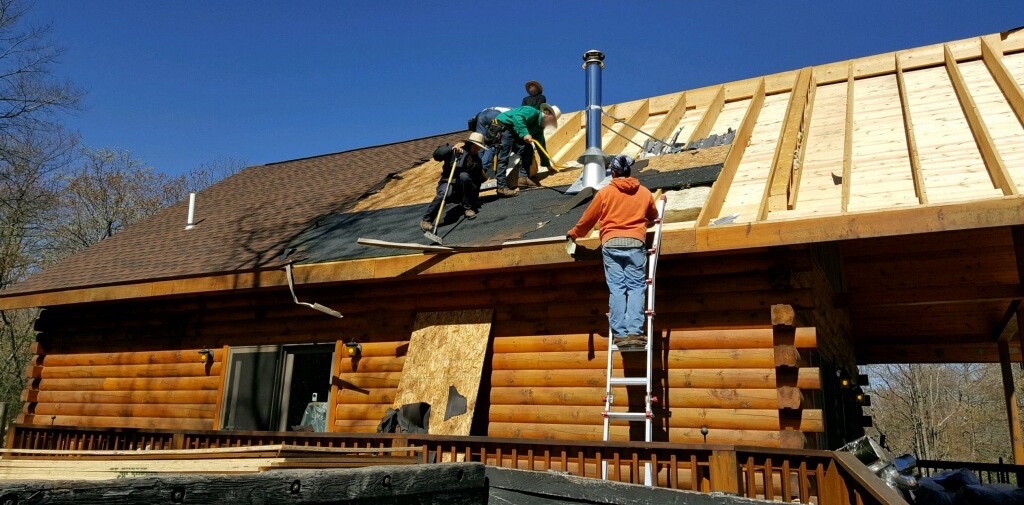 Three Amish men on the roof of a log cabin ripping up shingles with a fourth man in an orange sweat shirt standing on a ladder that is leaning against the side of the house.