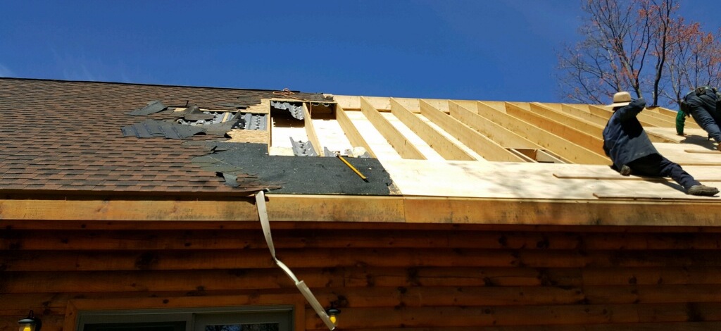 Two Amish men sitting on the roof of a log cabin that has half of the shingles ripped off exposing the wood underneath.