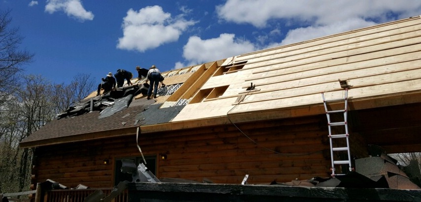 Four Amish men on the roof of a log cabin ripping off black roof shingles.