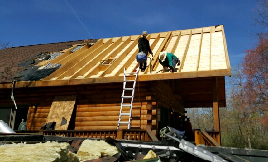 Two Amish men on the roof of a log cabin standing inside of the wooden bays after the shingles and plywood had been removed from that half of the roof. There is a ladder leaning against the side of the building.