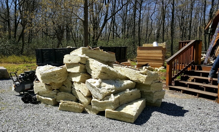 A very large pile of yellow fiberglass batts in the driveway in front of wooden log cabin steps. There is a pile of new plywood behind it.