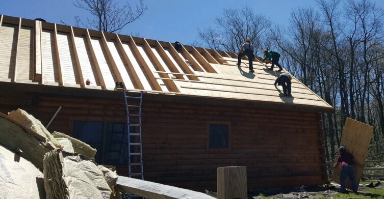 The side of a log cabin with the roof shingles taken off exposing the wood under. New plywood is being put down and there is a large pile of yellow fiberglass in front of the cabin on the ground. Three men are up on the roof.