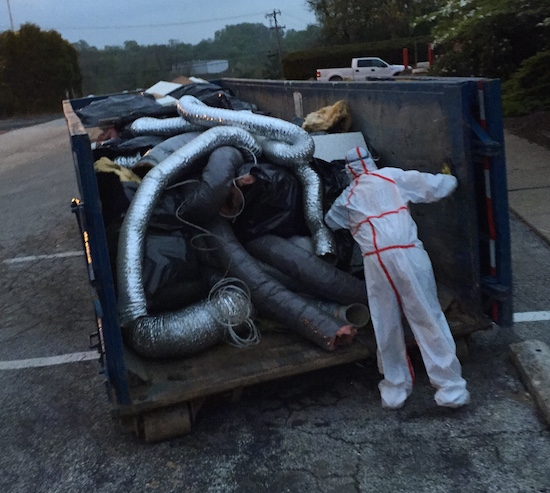 A man in a hazmat suit in front of a dumpster full of fiberglass lined duct work which was removed from a building being removed because of the hazards of fiberglass