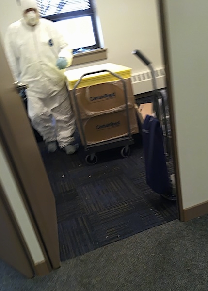 A man wearing a white hazmat suit, a face mask and gloves inside of an offfice standing on a blue carpet with a stack of fiberglass ceiling panels and a vacuum cleaner next to him.