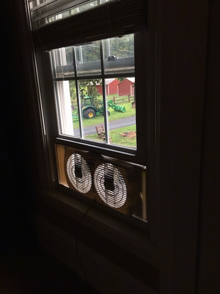 A fan in a window view from the inside of the home, with a green John Deere Tractor and red run-in shed barns out the window