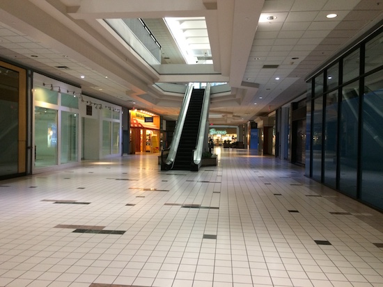 An empty shopping mall with an escalator and closed stores. There is a bright sky light letting in the sunlight.