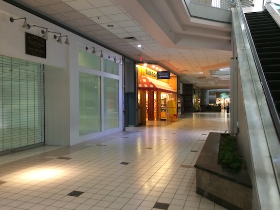 The isle in an empty shopping mall with closed stores, and a shiny white tiled floor. There is a bath soap store with a red and white awning and bright lights but no people.