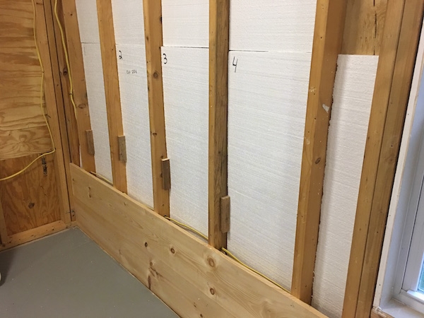 The studs behind a wall with a Styrofoam panels inbetween them with the first two wood boards from the wall across the bottom nailed in. The floor is painted gray.