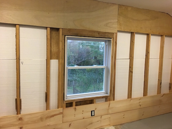 A wall of studs with Styrofoam panels inbetween them next to a window with the first two boards from the wall across the bottom nailed in. There is a cut out in the wood around the electrical outlet.